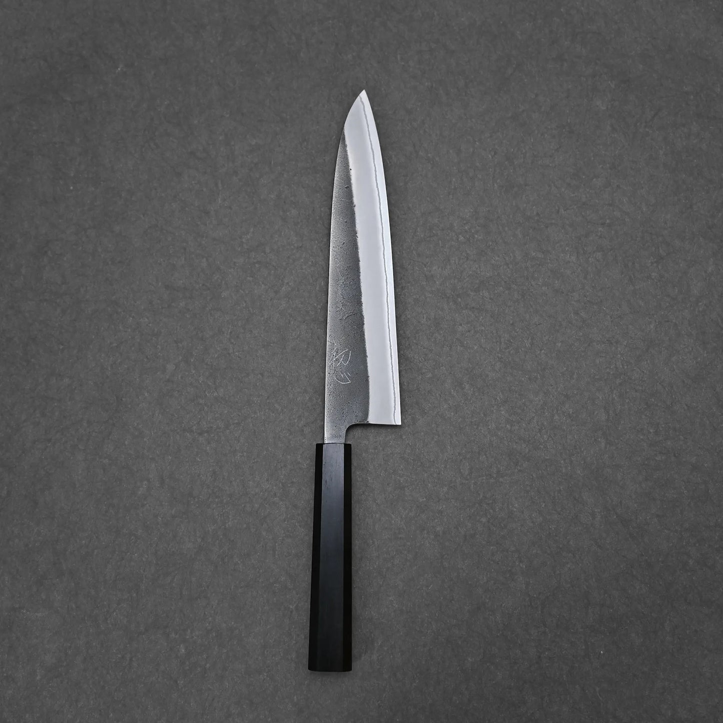A single Hatsukokoro x Yoshikane kurouchi nashiji Shirogami#2 gyuto 210mm chefs knife with a black handle is placed vertically, blade facing upward, on a plain dark grey surface.