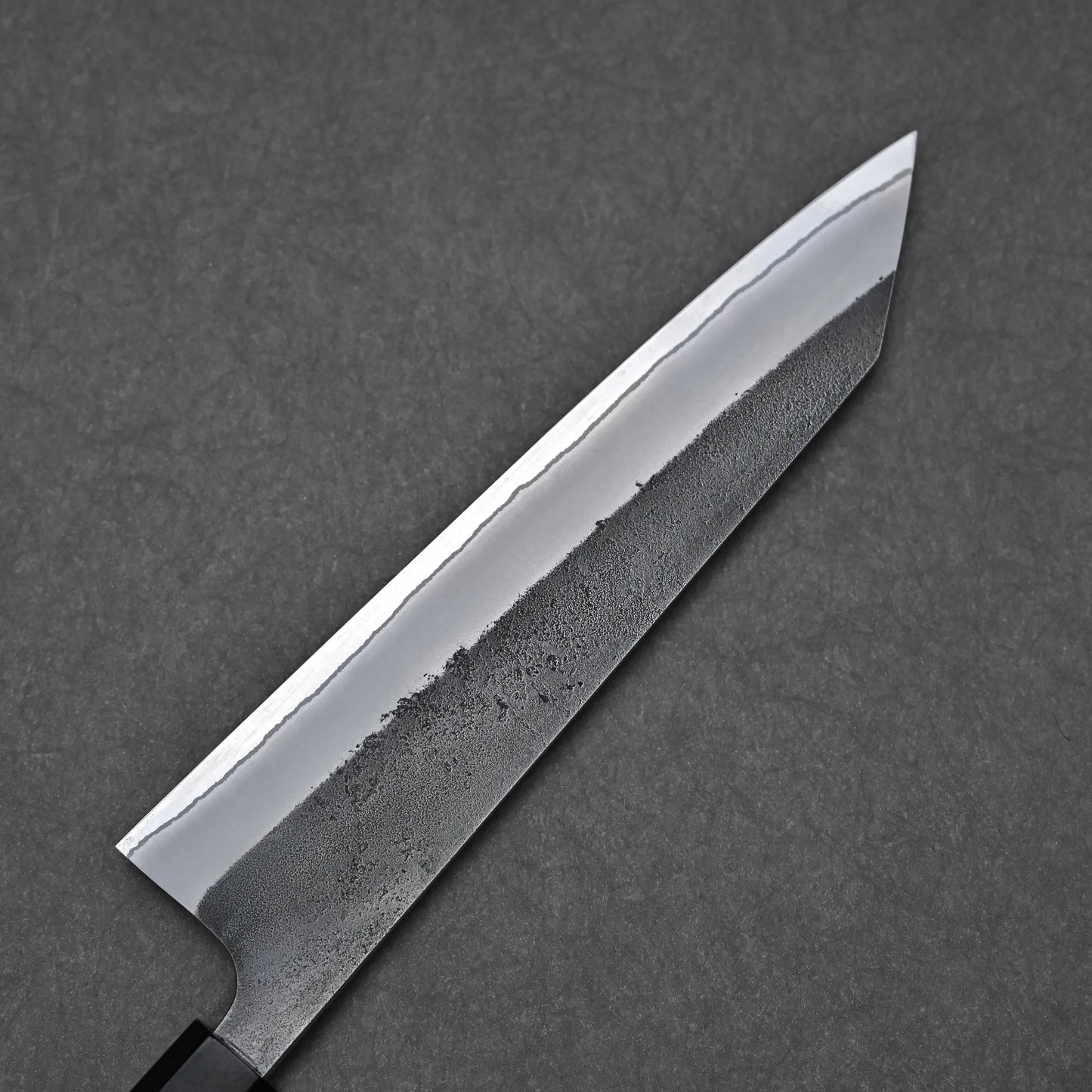 A close-up of the Hatsukokoro x Yoshikane kurouchi nashiji shirogami#2 kiritsuke gyuto 210mm, showing its sharp, shiny blade with a textured nashiji finish near the edge, resting on a dark, textured background.