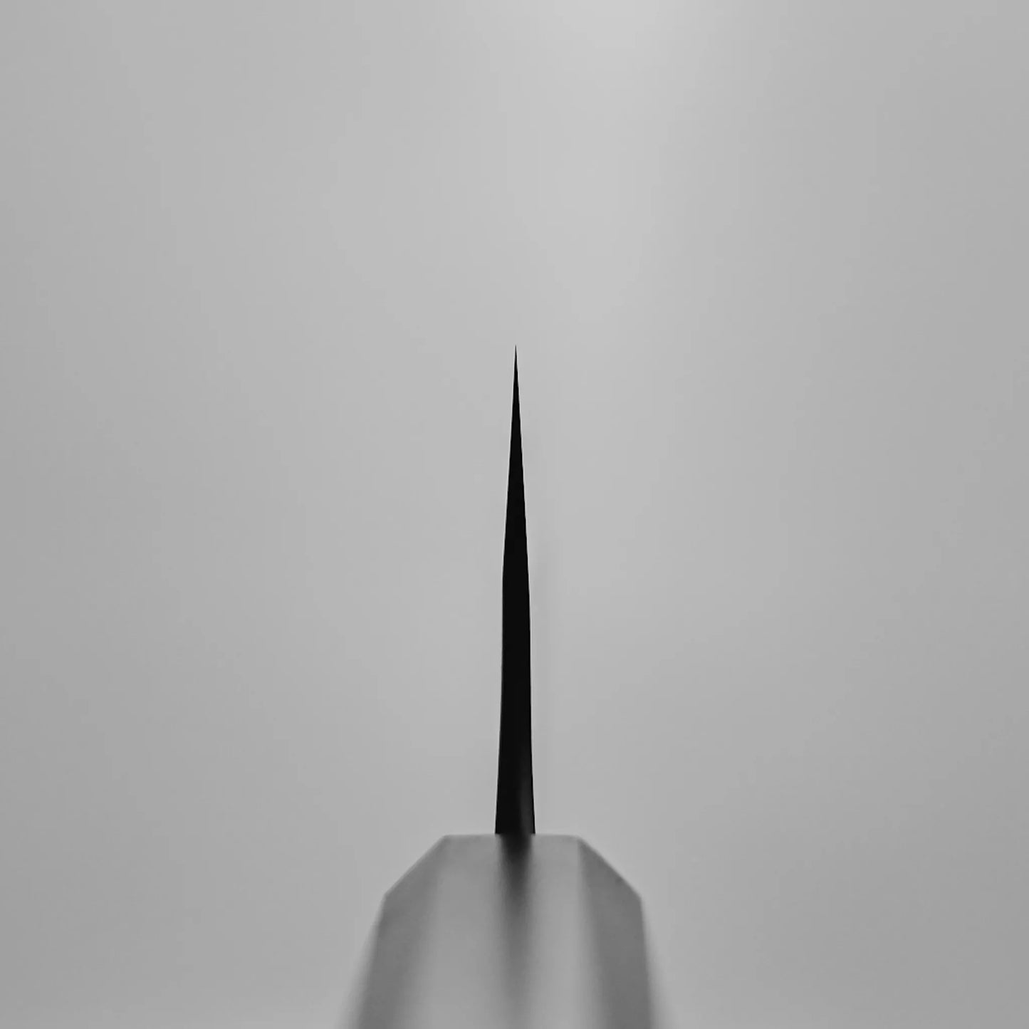Close-up, front-facing view of the thin, pointed edge of a Hatsukokoro x Yoshikane kurouchi nashiji shirogami#2 kiritsuke gyuto 210mm, its sharp blade vertical against a plain light gray background.