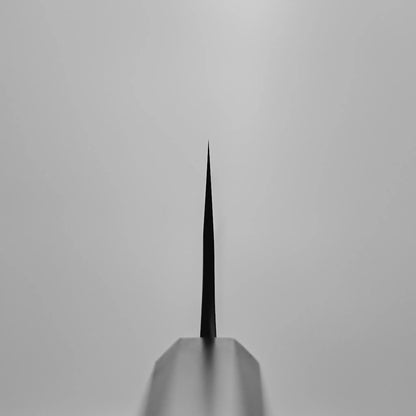 Close-up, front-facing view of the thin, pointed edge of a Hatsukokoro x Yoshikane kurouchi nashiji shirogami#2 kiritsuke gyuto 210mm, its sharp blade vertical against a plain light gray background.