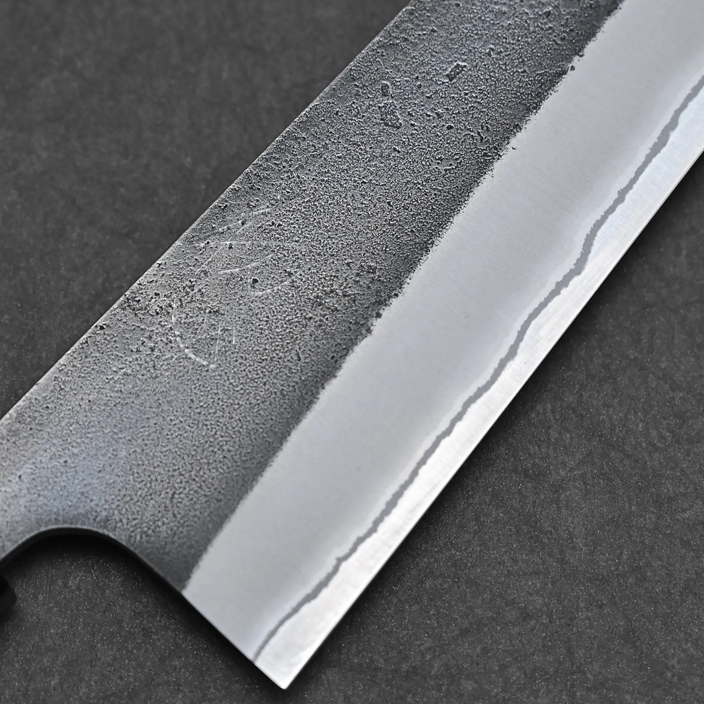 Close-up of a Hatsukokoro x Yoshikane kurouchi nashiji shirogami#2 kiritsuke gyuto 210mm blade, highlighting its textured matte upper section, polished edge, and wavy hamon against a dark background.