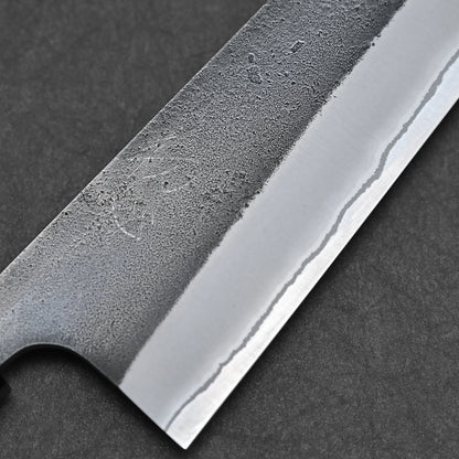 Close-up of a Hatsukokoro x Yoshikane kurouchi nashiji shirogami#2 kiritsuke gyuto 210mm blade, highlighting its textured matte upper section, polished edge, and wavy hamon against a dark background.