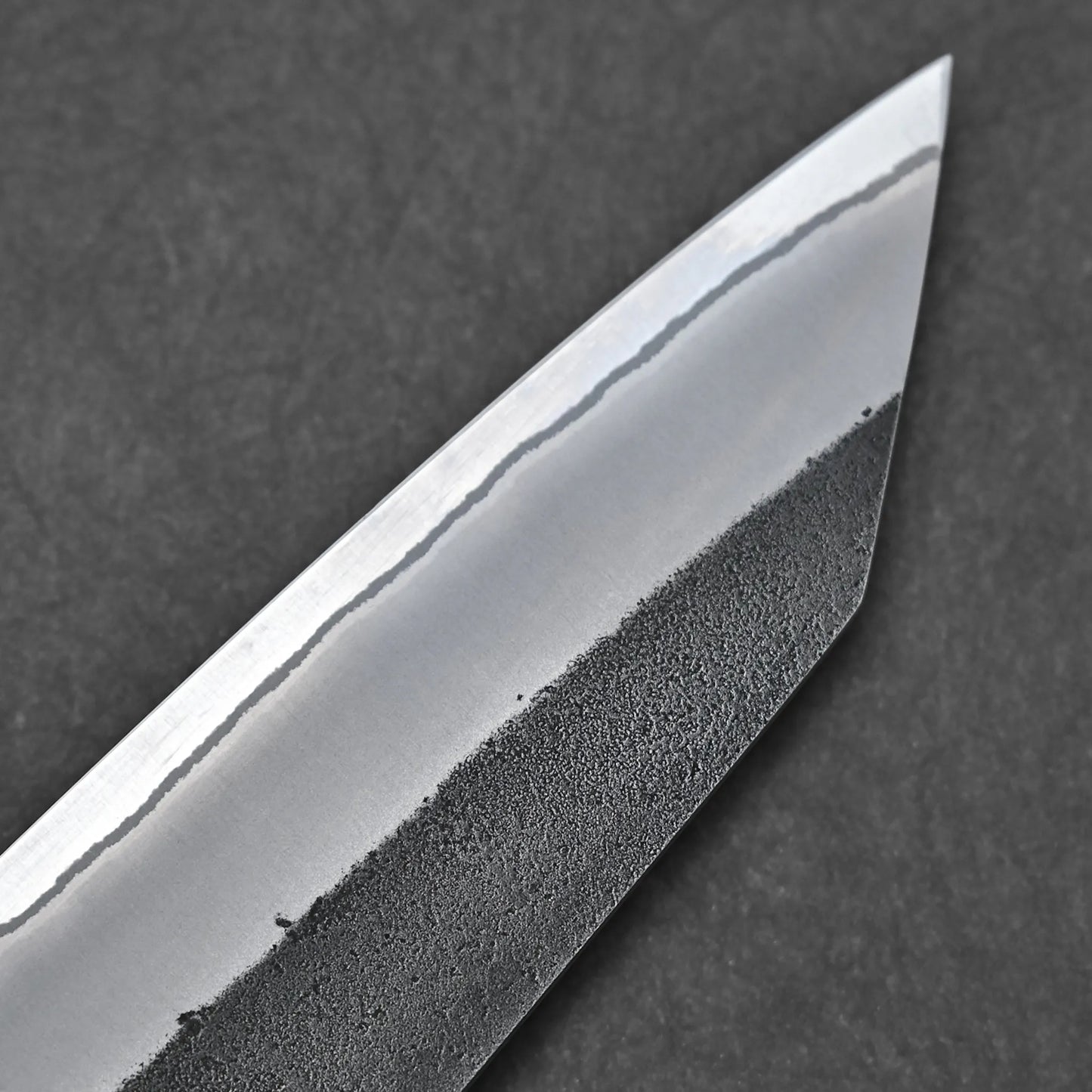Close-up of the Hatsukokoro x Yoshikane kurouchi nashiji shirogami#2 kiritsuke gyuto 210mm blade tip, highlighting its distinct hamon line and polished edge contrasting with the rustic spine, set against a dark background.