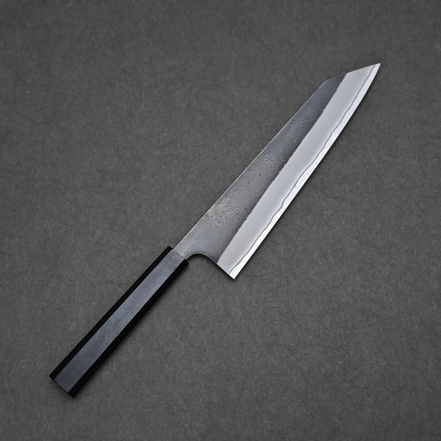 A Hatsukokoro x Yoshikane kurouchi nashiji shirogami#2 kiritsuke gyuto 210mm with a rectangular handle and straight-edged pointed blade rests on a dark, textured surface.