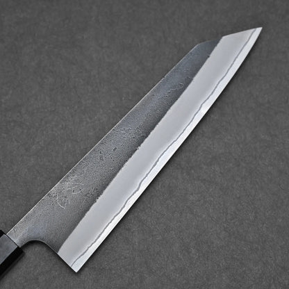 A close-up of the Hatsukokoro x Yoshikane kurouchi nashiji shirogami#2 kiritsuke gyuto 210mm, featuring a sharp pointed blade with a wavy pattern, set against a dark gray textured background.