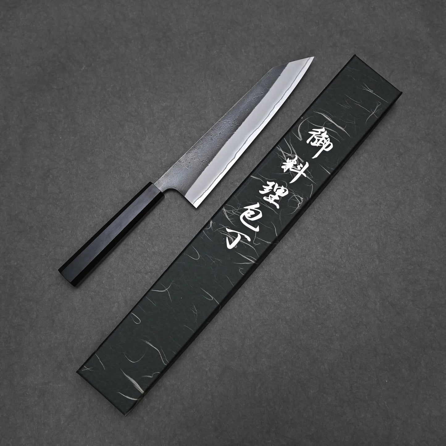 A Hatsukokoro x Yoshikane kurouchi nashiji shirogami#2 kiritsuke gyuto 210mm with a black handle is displayed next to a rectangular black case featuring white Japanese characters, both set on a dark textured surface.
