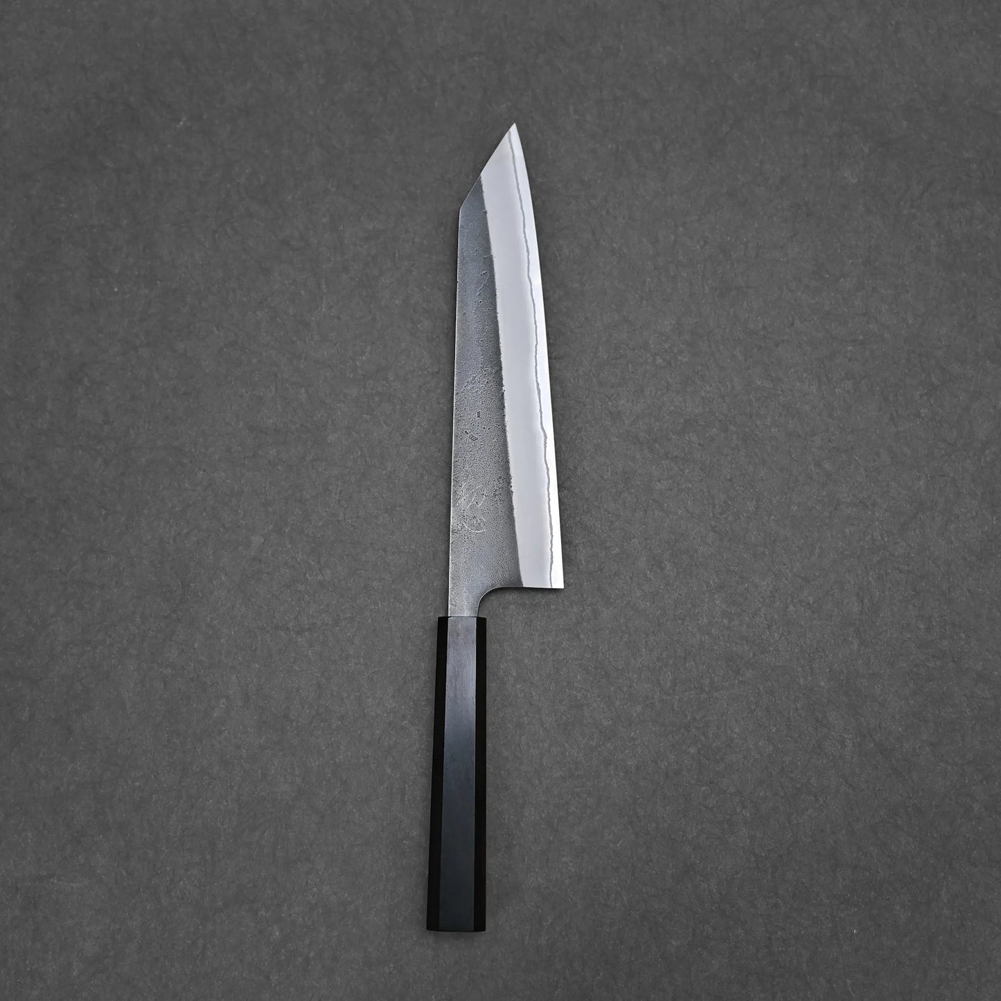 The Hatsukokoro x Yoshikane kurouchi nashiji shirogami#2 kiritsuke gyuto 210mm, featuring a straight-edged carbon steel blade and black rectangular handle, is placed on a gray surface.