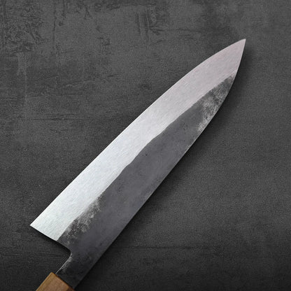 A gyuto with a dark handle is displayed; the blade has a tall profile and subtle texture.