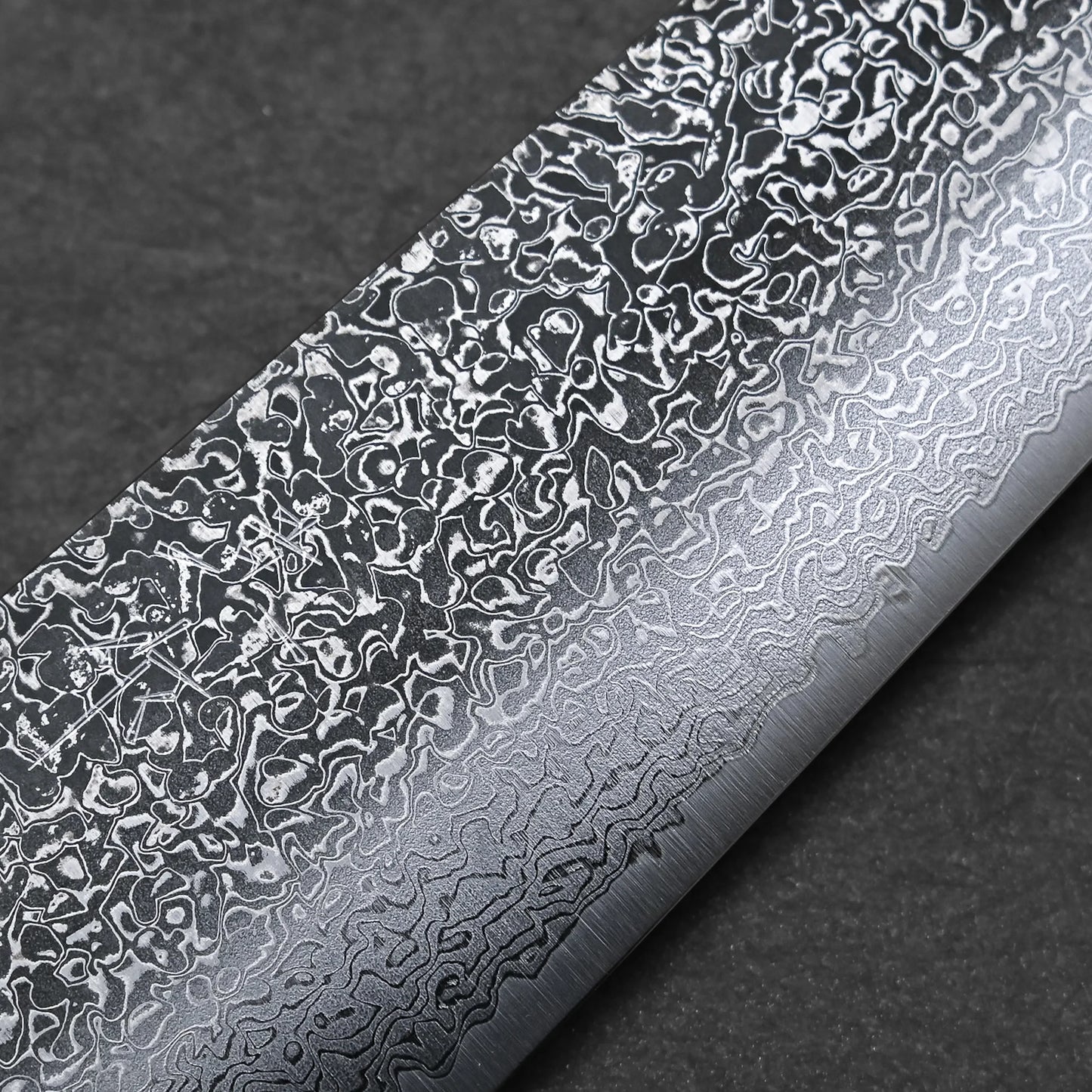Close-up of the Kei Kobayashi SG2 damascus gyuto 240mm blade, highlighting intricate wavy and mottled patterns on the metal surface, set against a dark, textured background.