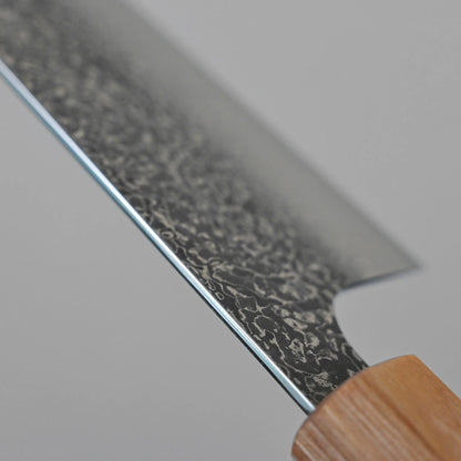 Close-up of the Kei Kobayashi SG2 damascus gyuto 240mm kitchen knife, showing its wooden handle and dark, patterned blade with a textured finish, set against a plain grey background.
