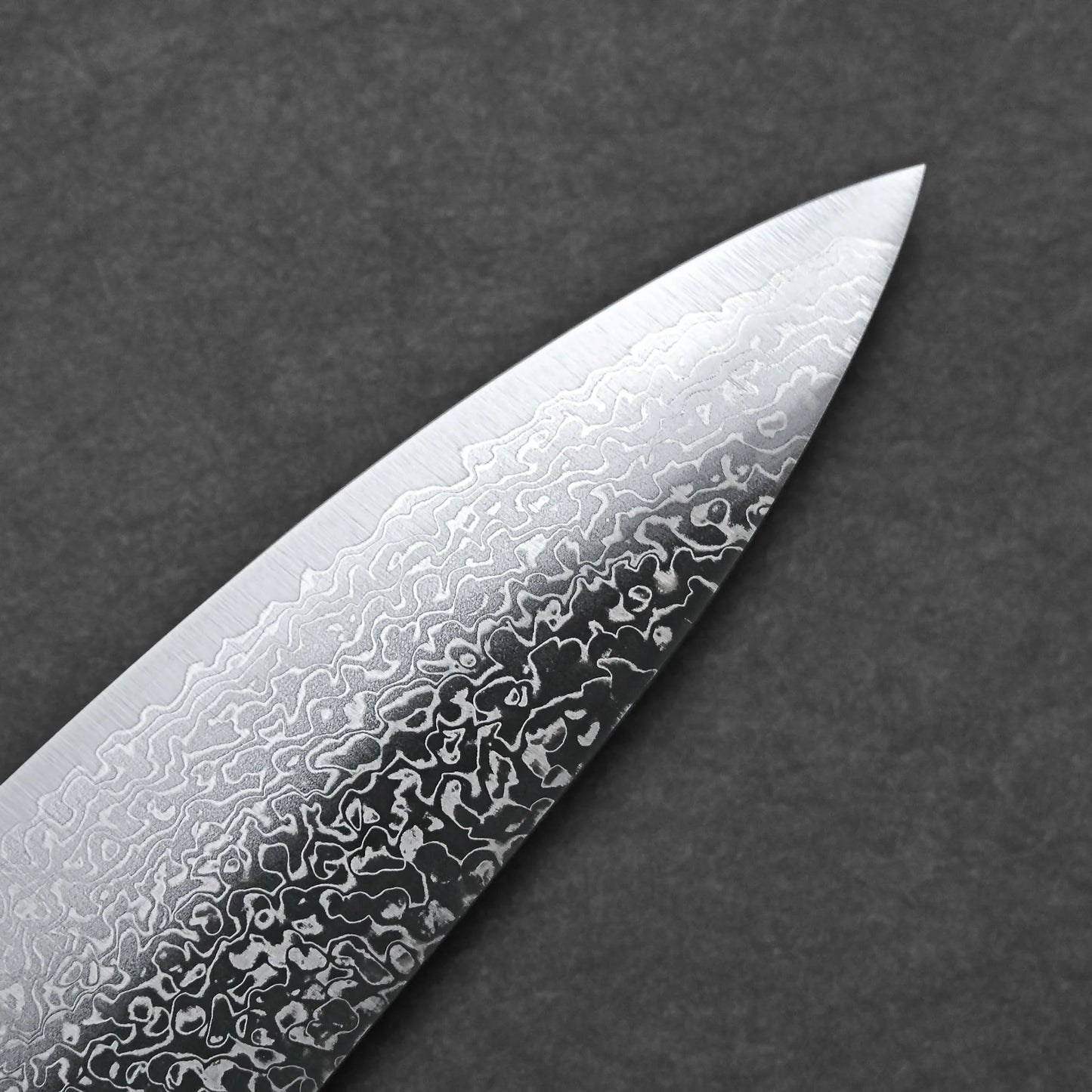 Close-up of the tip of the Kei Kobayashi SG2 damascus gyuto 240mm kitchen knife blade, showing its wavy, patterned texture against a plain dark gray background.