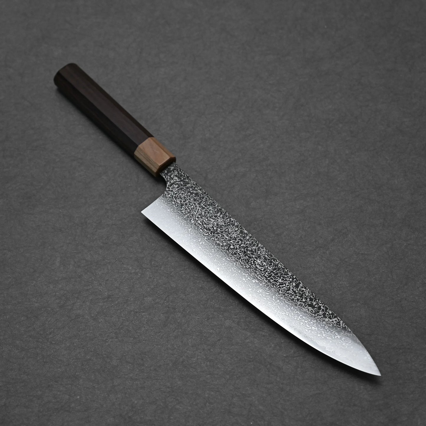 The Kei Kobayashi SG2 damascus gyuto 240mm, featuring a textured, speckled blade and dark wooden handle, rests on a dark gray surface to showcase the elegance of traditional Japanese craftsmanship.
