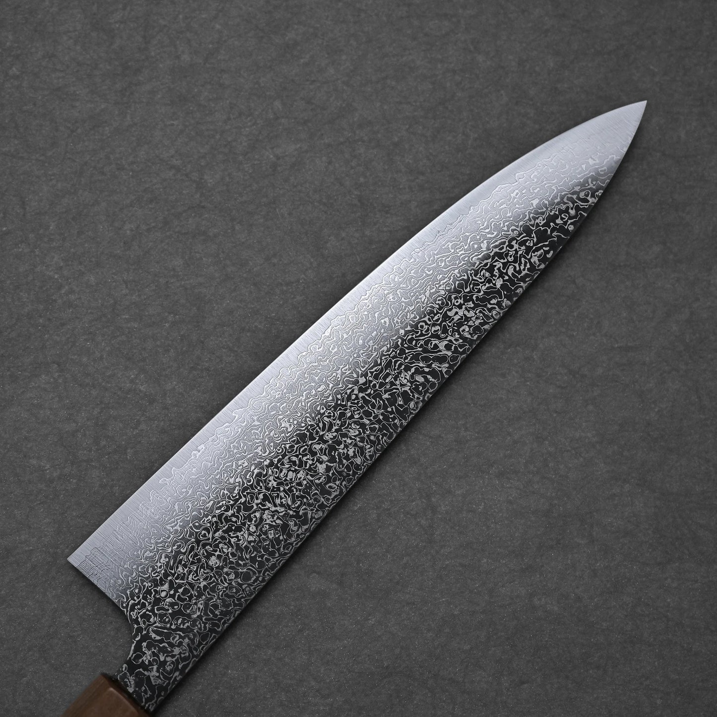 A close-up of the Kei Kobayashi SG2 damascus gyuto 240mm showcases intricate swirling patterns on its blade against a dark textured background, with its handle partially visible in the lower left corner.
