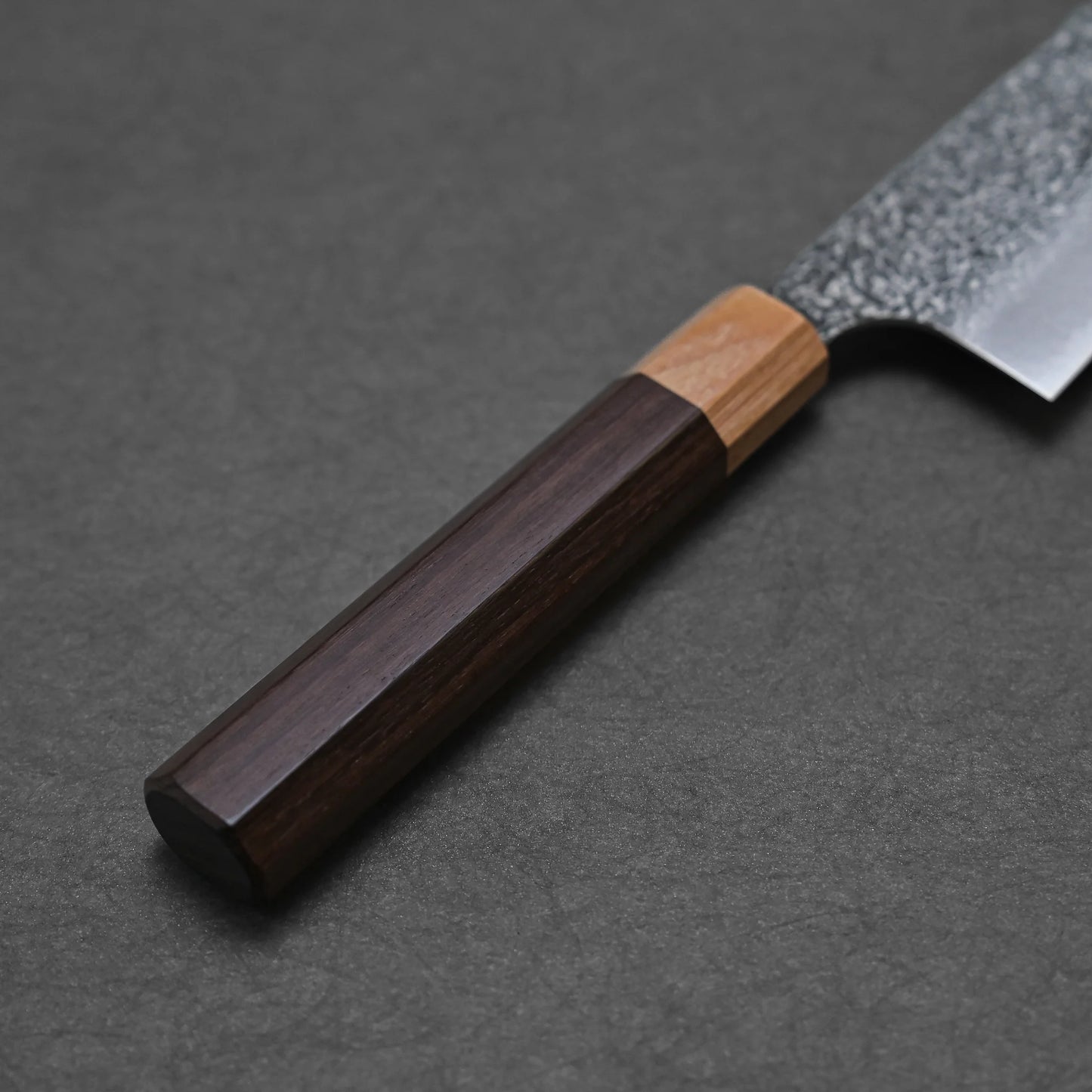A close-up shows the dark and light wood handle of the Kei Kobayashi SG2 damascus gyuto 240mm resting on a dark textured surface, with part of its blade visible in the top right corner.