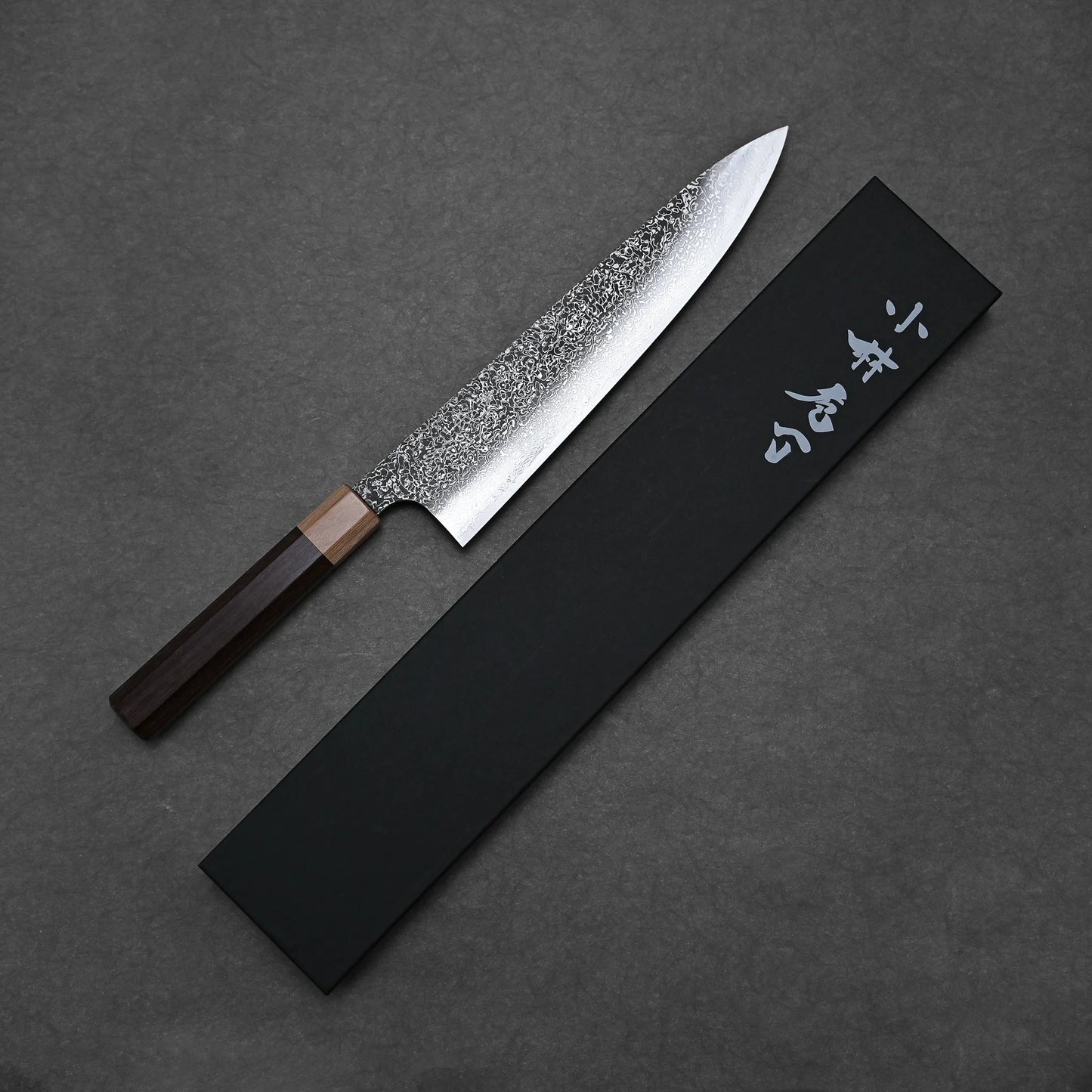 The Kei Kobayashi SG2 damascus gyuto 240mm, featuring a textured blade and wooden handle, rests beside a black box with white Japanese characters on a dark grey background.