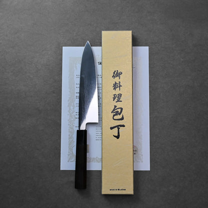 A Mizuhonyaki Japanese knife, gyuto type, with a curved blade and dark handle lying next to a beige box with certificate.