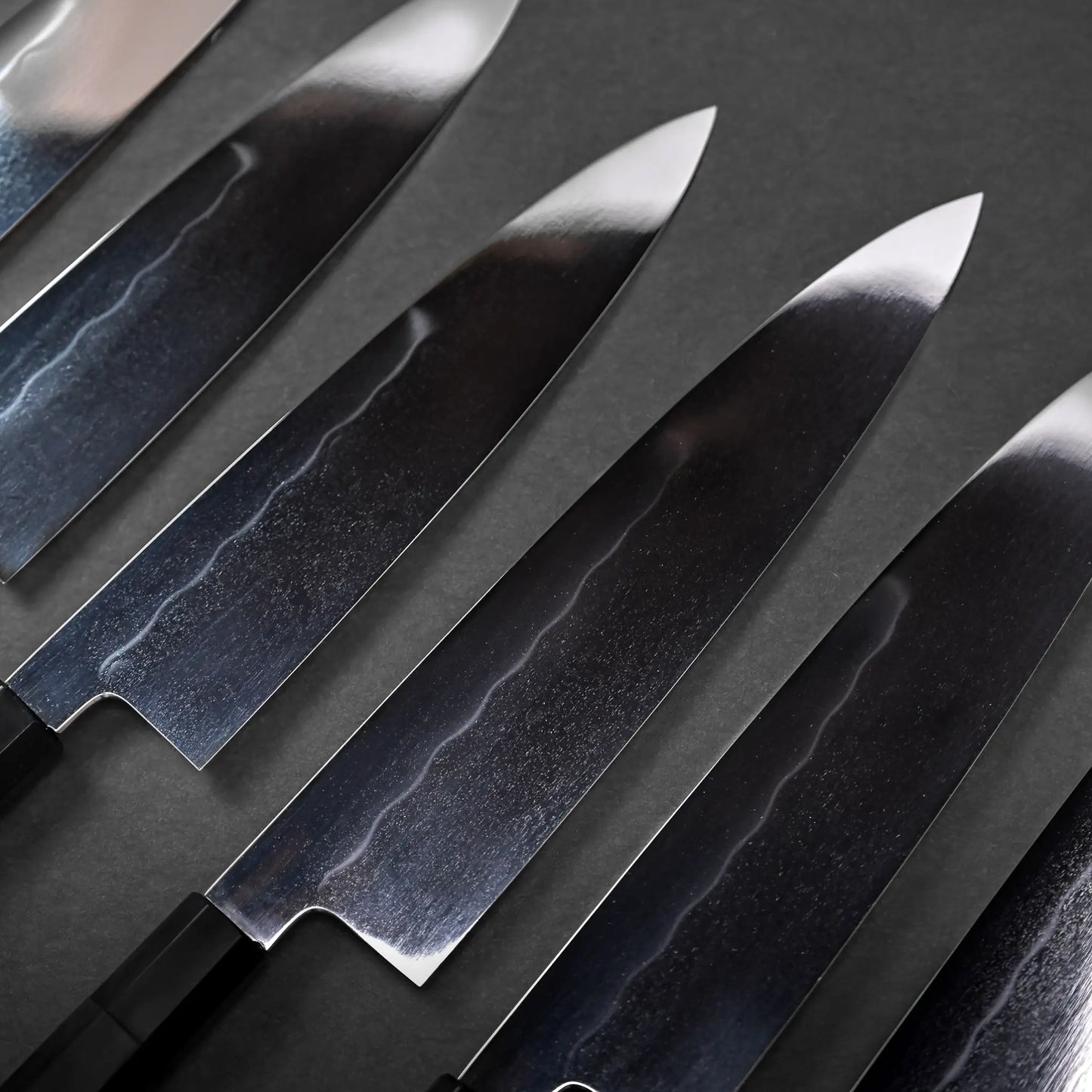 A row of gyuto blades with a mirrored finish, Japanese knife handles not visible, showing classic chef's knife shapes.