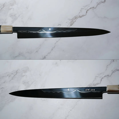 Two views of a Japanese honyaki yanagiba knife placed on a marble-patterned surface. The long, single-bevel blade has a pointed tip and features a decorative hamon resembling Mt. Fuji, indicating differential hardening. The top view shows a circular moon mark near the spine; the bottom view reveals a crescent moon motif and engraved kanji near the handle.
