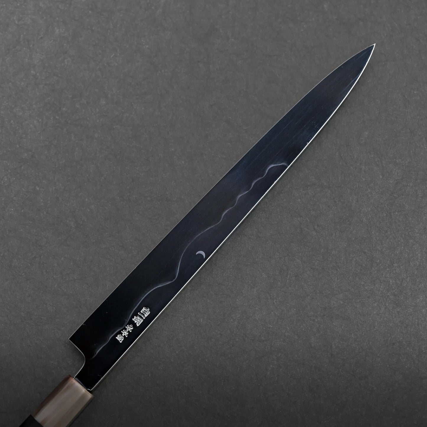 A mizuhonyaki yanagiba, a Japanese kitchen knife with a long, narrow blade and wooden handle, rests on a dark, textured surface. Japanese characters are engraved near the base of this honyaki sushi knife.