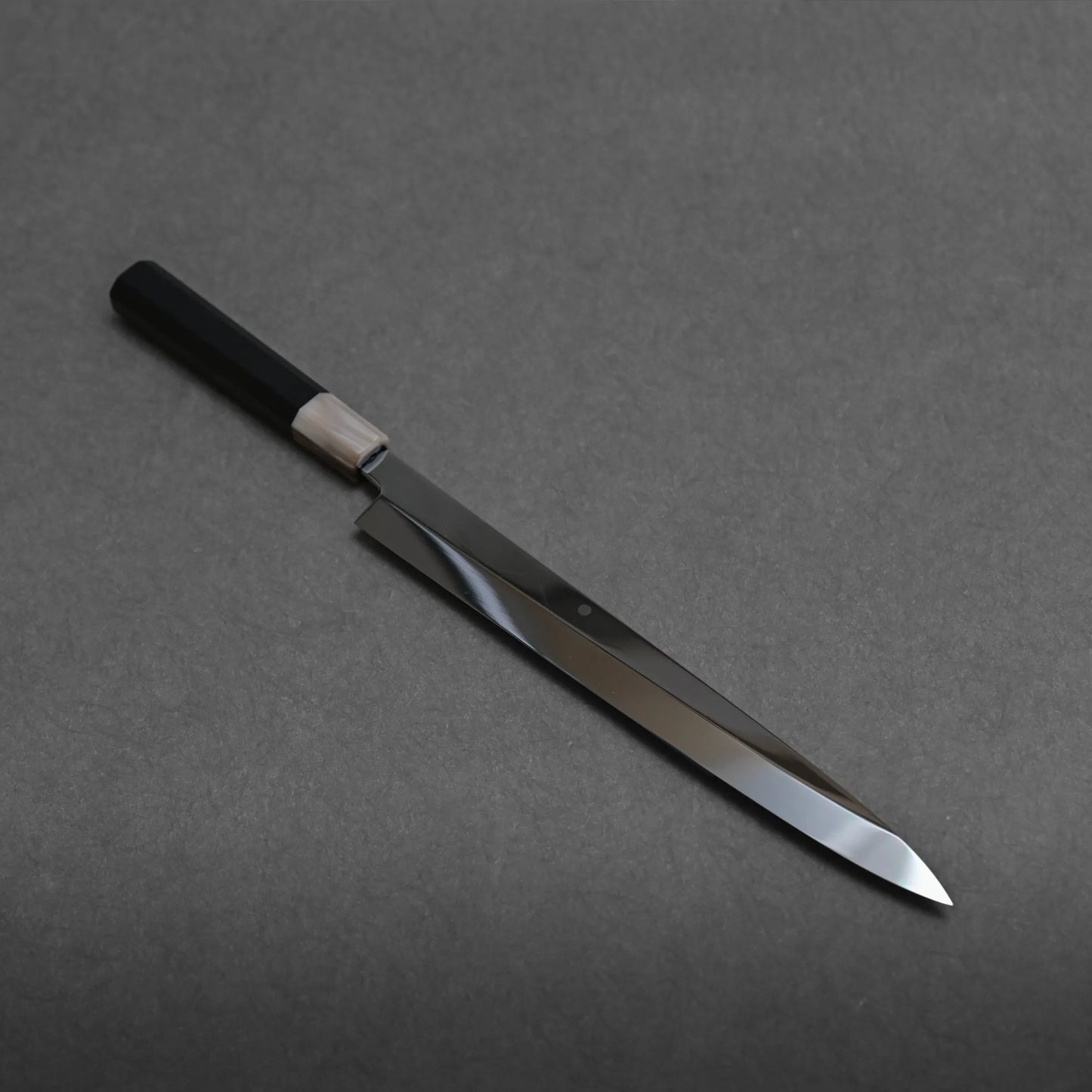 A Japanese-style mizuhonyaki yanagiba kitchen knife with a black handle and a long, narrow, shiny blade is placed diagonally on a plain dark gray surface.