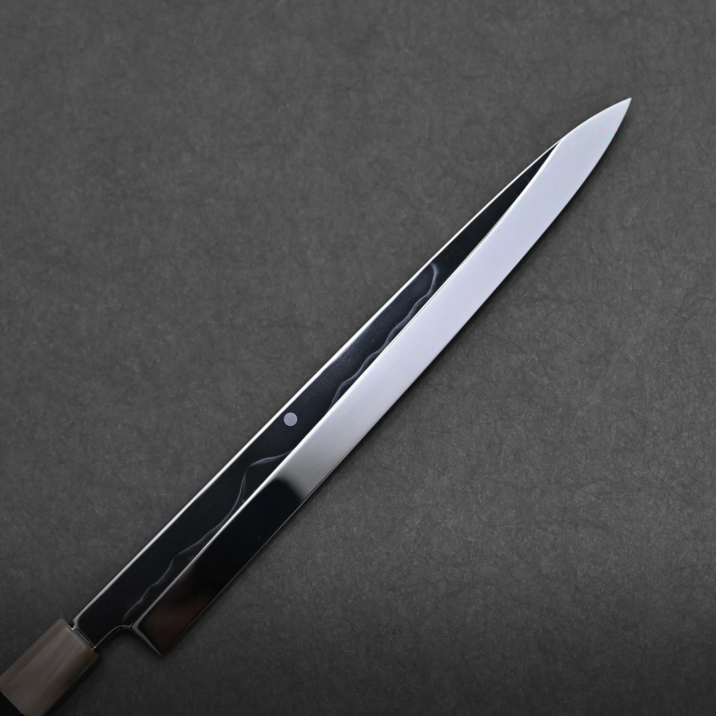 A close-up view of a polished mizuhonyaki yanagiba, a single-edged Japanese honyaki sushi knife with a sharp, pointed blade against a dark, textured background.