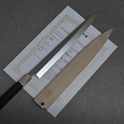 A long, single-edged mizuhonyaki yanagiba with a black handle lies on a document with Japanese text. Beside it rests a wooden sheath. Both the honyaki sushi knife and sheath are set on a dark gray surface.