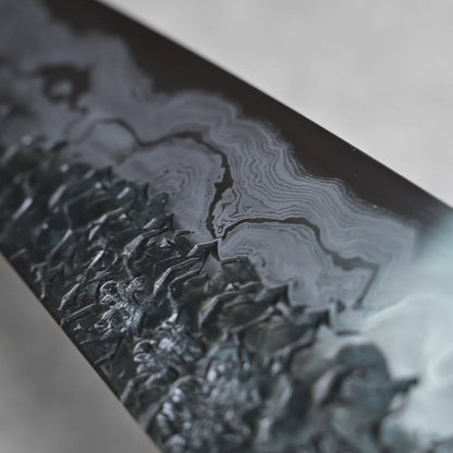 Close-up angled view of a gyuto knife blade showing a detailed Damascus pattern. The lower section has a dark, hammered texture that shifts into lighter, flowing wavy layers near the spine.