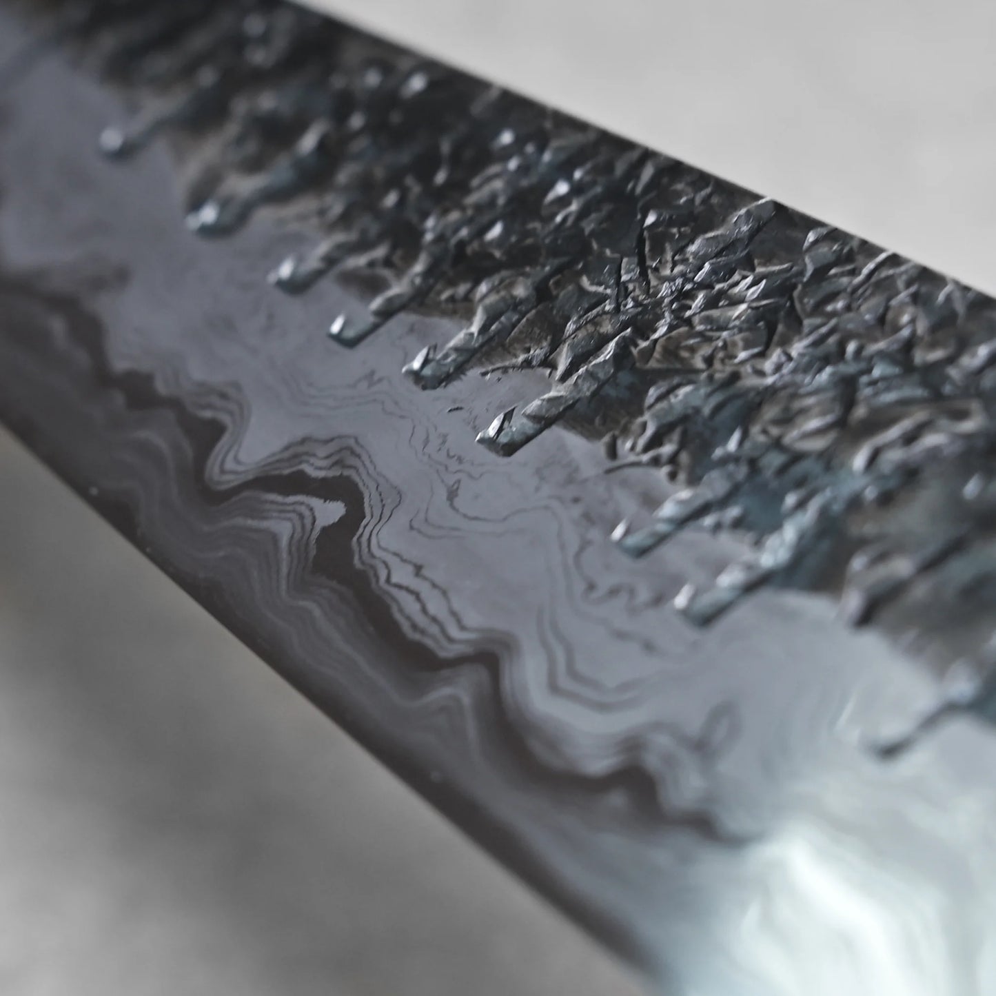 Close-up angled view of the reverse side of a gyuto knife blade. The lower area has a dark, hammered texture that transitions into lighter, wavy layers extending toward the spine.