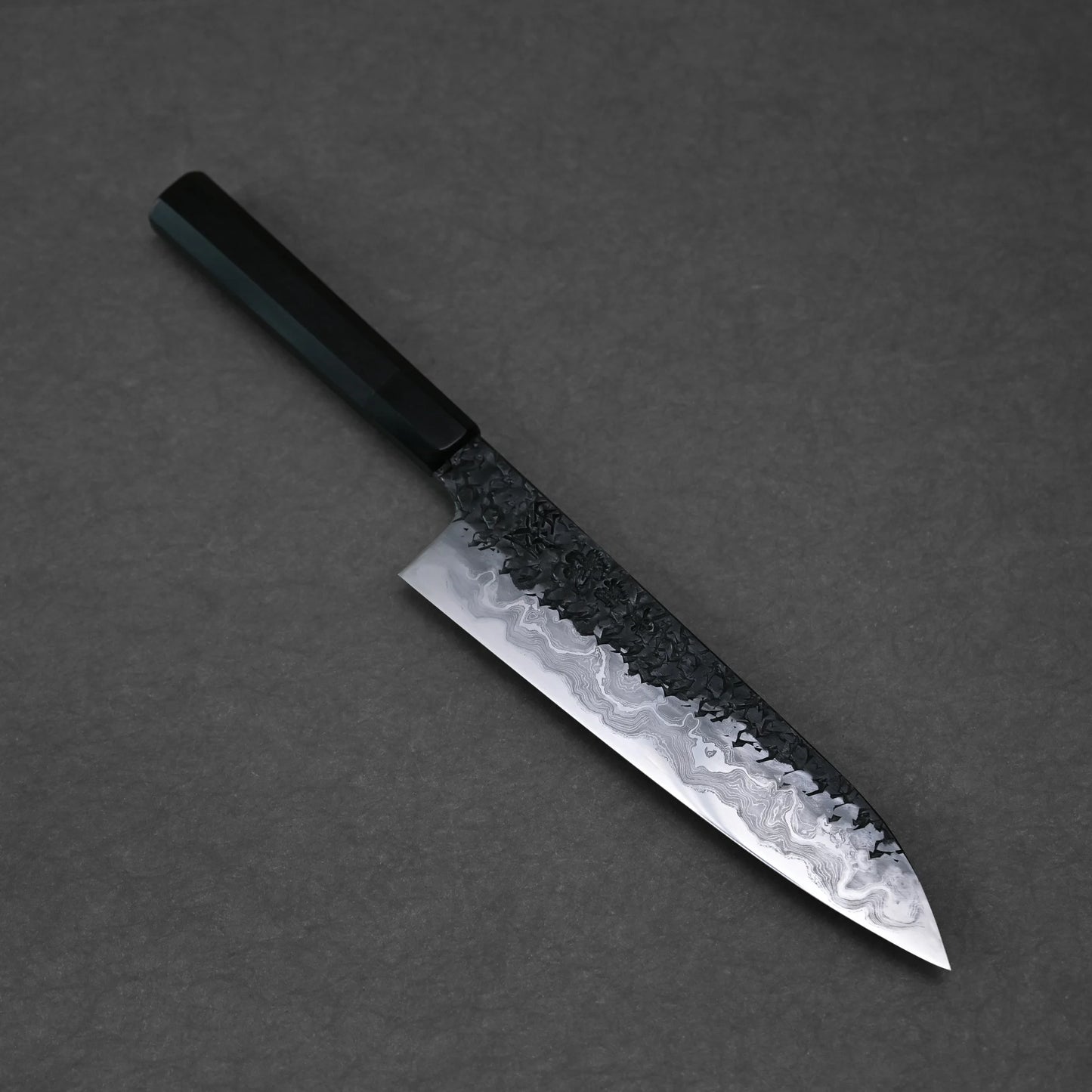 Overhead view of a diagonally positioned gyuto knife on a dark grey surface. The blade shows a Damascus pattern with a dark, hammered top and lighter, wavy lower section. The knife has a dark, octagonal handle.