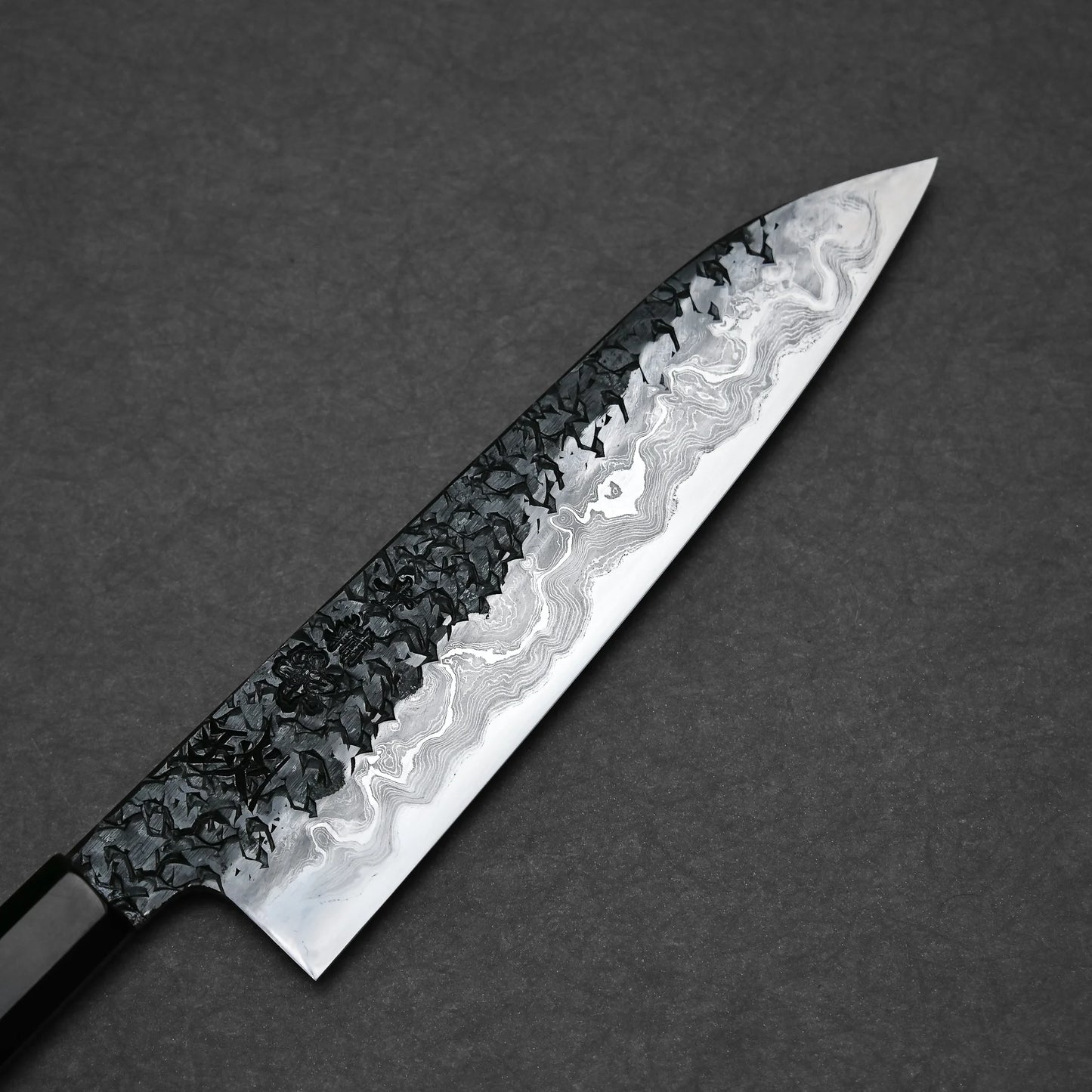 Close-up of a gyuto knife blade with an intricate Damascus pattern. The top section has a dark, hammered texture, transitioning into lighter, wavy layers toward the sharpened edge. The blade tapers to a pointed tip.