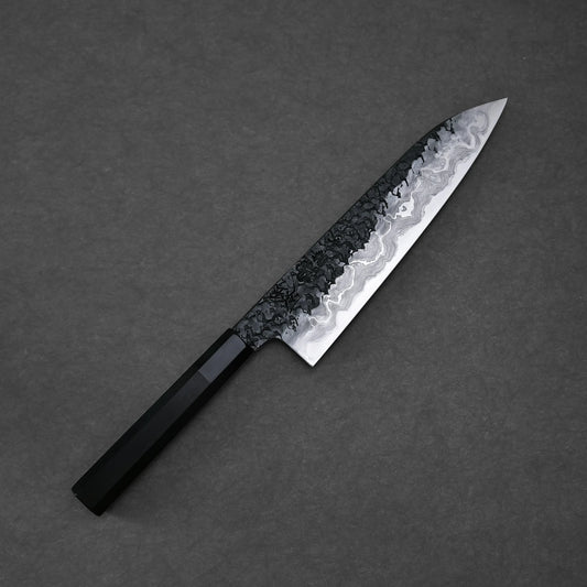 Diagonal view of a gyuto knife on a textured grey surface. The blade has a Damascus pattern with dark, hammered details on the upper section and lighter, wavy layers on the polished lower half. The handle is dark and octagonal.