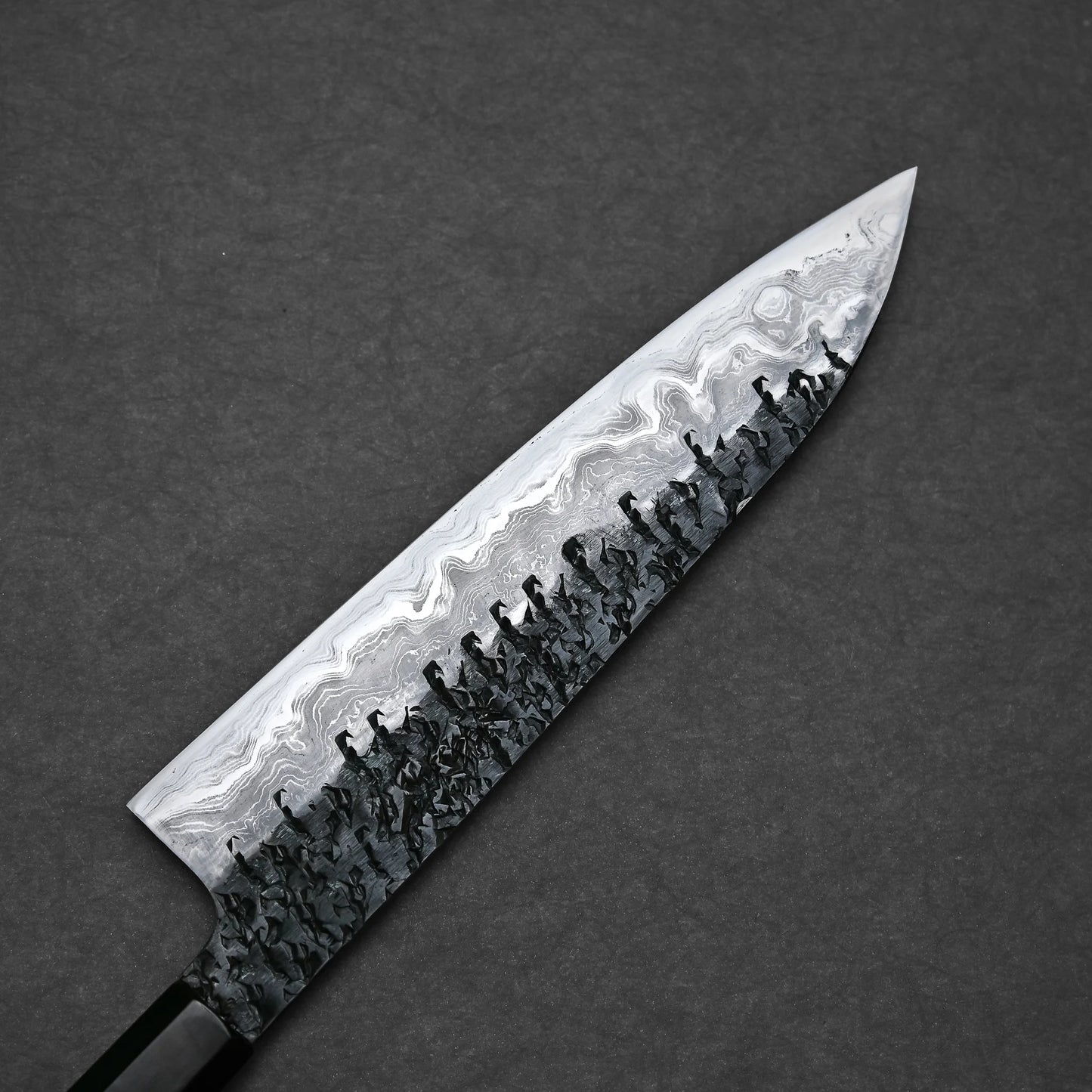 Close-up of the reverse side of a gyuto knife blade with a pronounced Damascus pattern. The lower portion has a dark, hammered texture that fades into lighter, wavy layers reaching up to the spine and pointed tip.