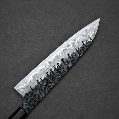Close-up of the reverse side of a gyuto knife blade with a pronounced Damascus pattern. The lower portion has a dark, hammered texture that fades into lighter, wavy layers reaching up to the spine and pointed tip.