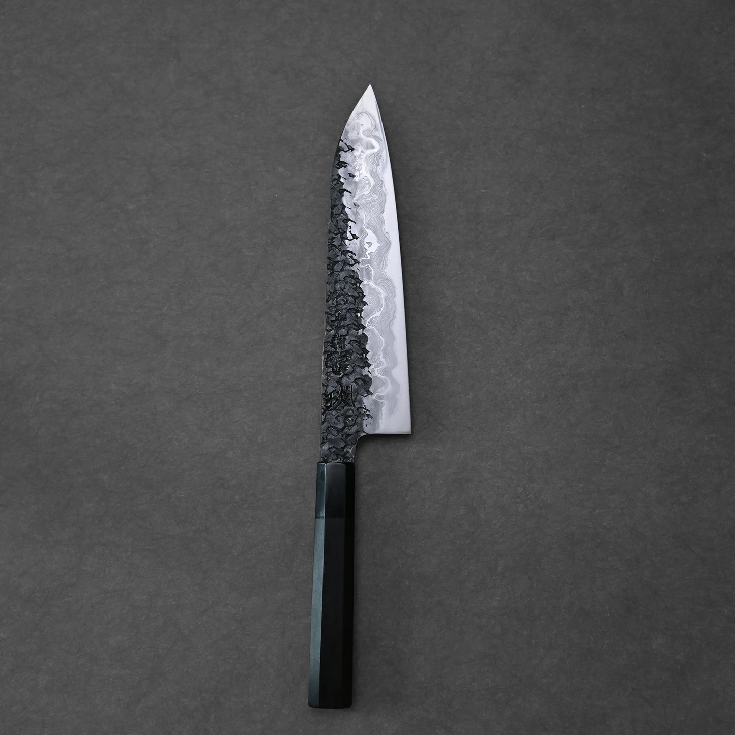 Top-down view of a vertically positioned gyuto knife on a dark grey surface. The blade features a bold Damascus pattern with a dark, hammered texture near the spine that fades into lighter, wavy layers. The knife has a dark, octagonal handle at the base.