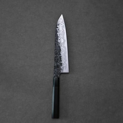 Top-down view of a vertically positioned gyuto knife on a dark grey surface. The blade features a bold Damascus pattern with a dark, hammered texture near the spine that fades into lighter, wavy layers. The knife has a dark, octagonal handle at the base.