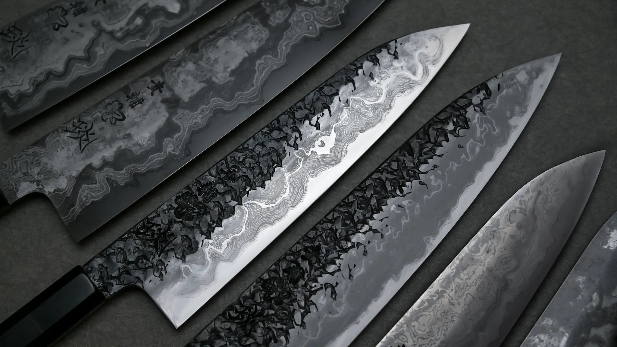 Japanese chef knives by Kisuke Manaka from the ENN series shown side by side. The blades have damascus patterns with strong contrast between steel layers.