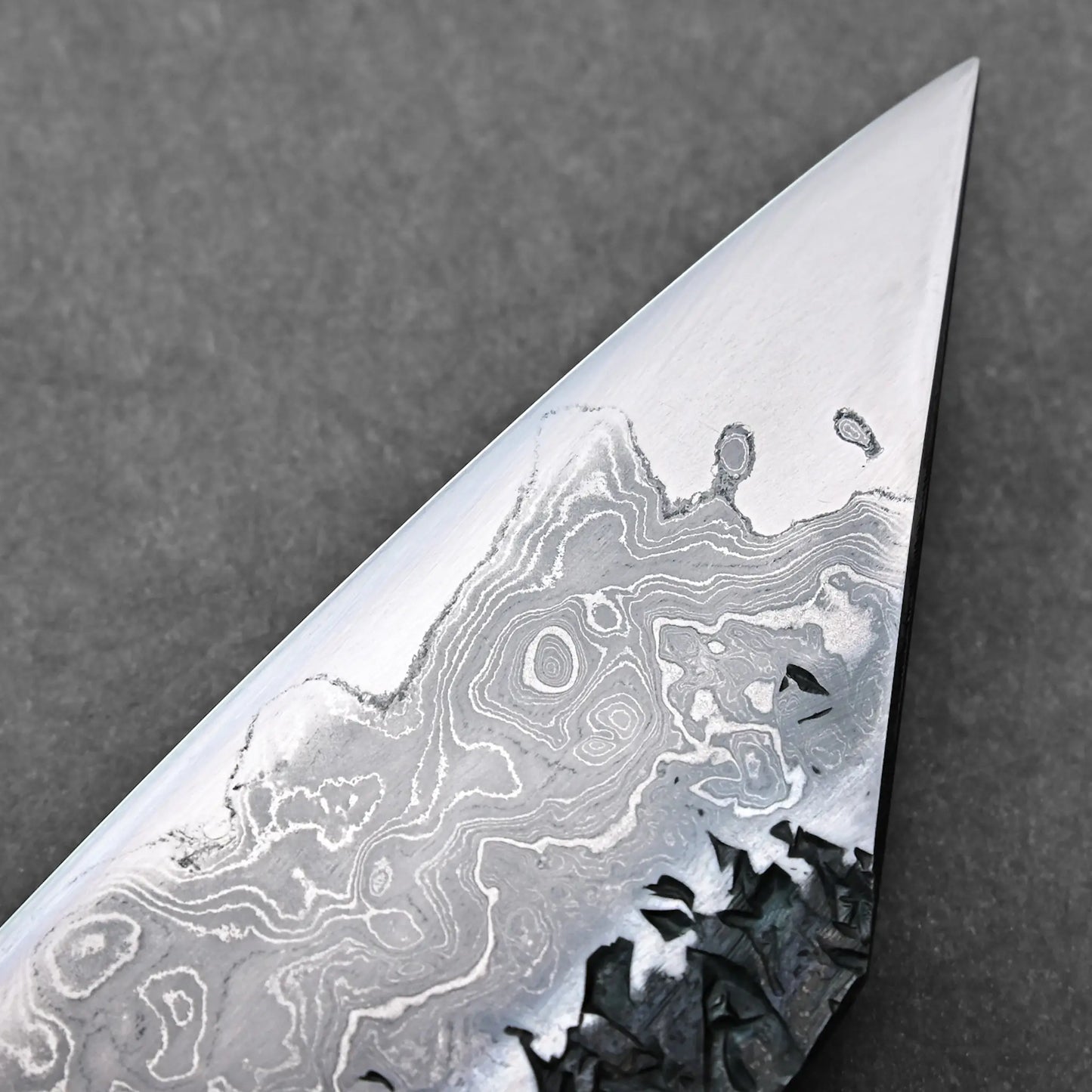 The image shows a close-up of a Japanese knife blade with visible damascus steel's wavy patterns and no handle in view.