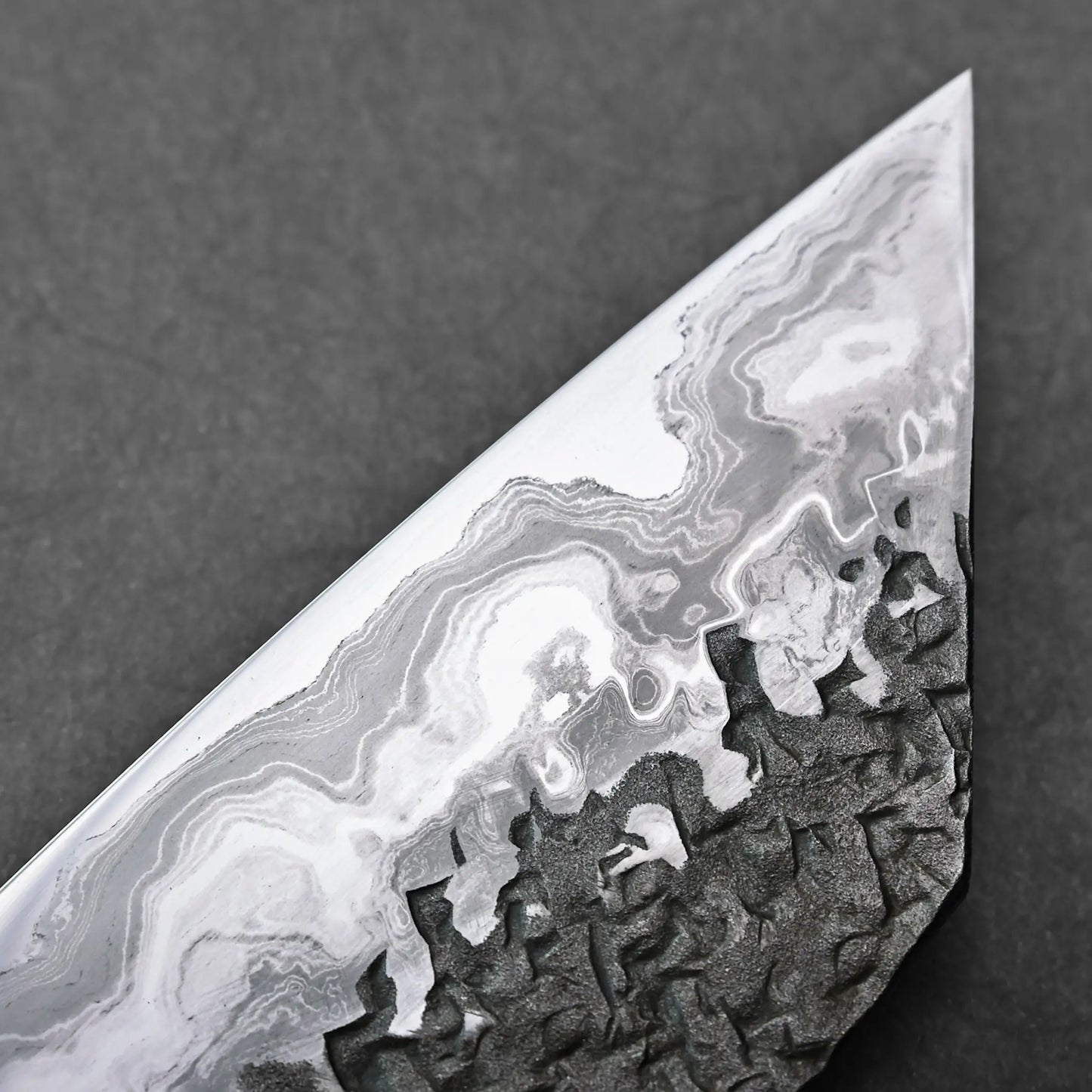 Close-up showing a Japanese knife blade with wavy Aogami #1 damascus layers and a dark, textured lower section.