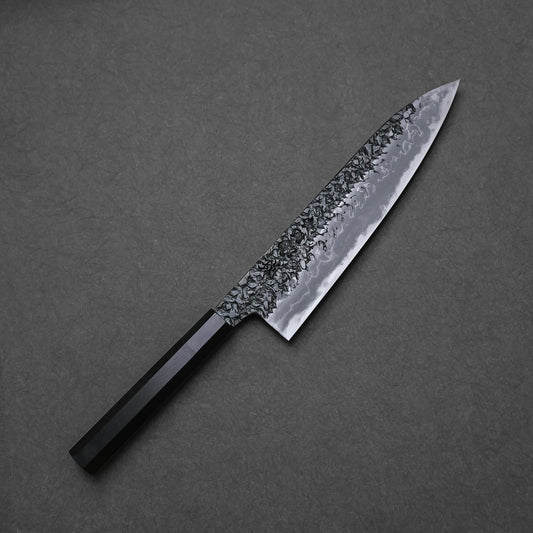 Japanese chef’s knife with tsuchime kurouchi damascus finish, featuring hammered texture, dark forged surface, and layered steel wave patterns; full blade photographed on dark background.