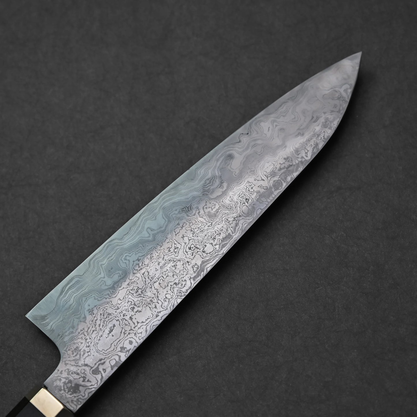 Reverse side of a Japanese Damascus steel knife blade with layered forge patterns and pointed tip, photographed against a dark textured background.