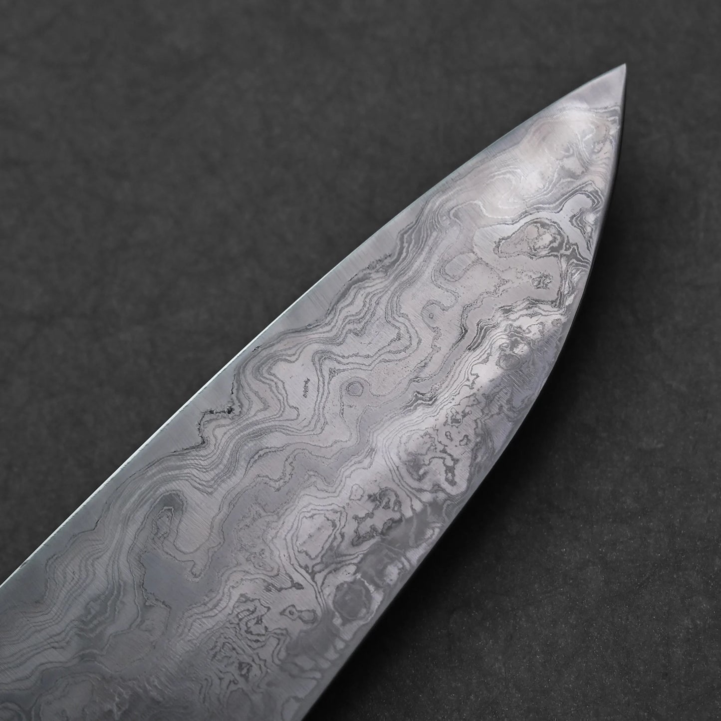 Reverse side of the tip of a Japanese Damascus steel knife blade showing fine layered forge patterns and swirling textures; photographed close-up against a dark background.