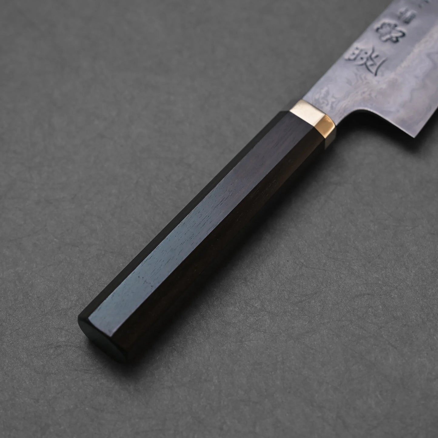 Japanese knife handle made of dark polished wood with a octagonal shape and brass bolster; adjacent blade section features vertically engraved kanji and a delicate floral motif.
