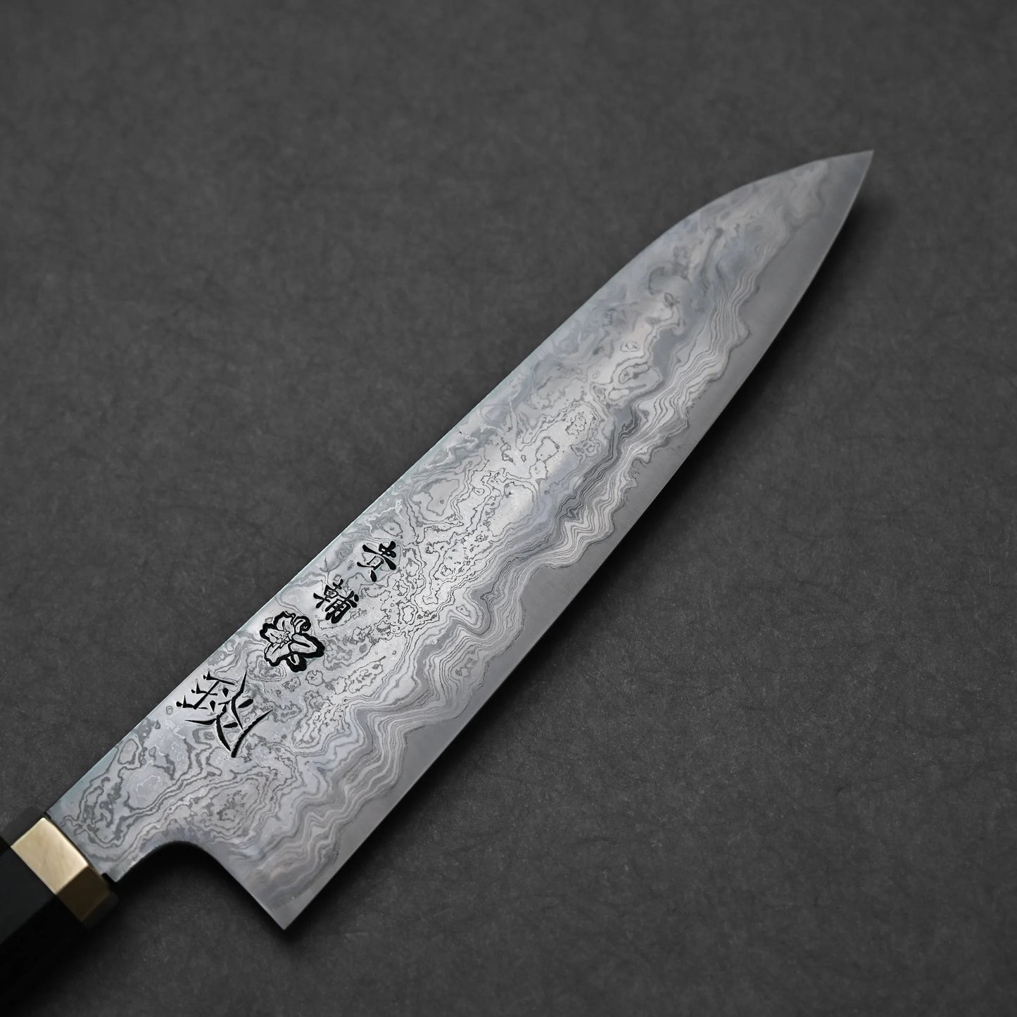Japanese Damascus steel knife blade with bold, swirling forge patterns and vertically engraved kanji characters; photographed in angled close-up against a textured surface.