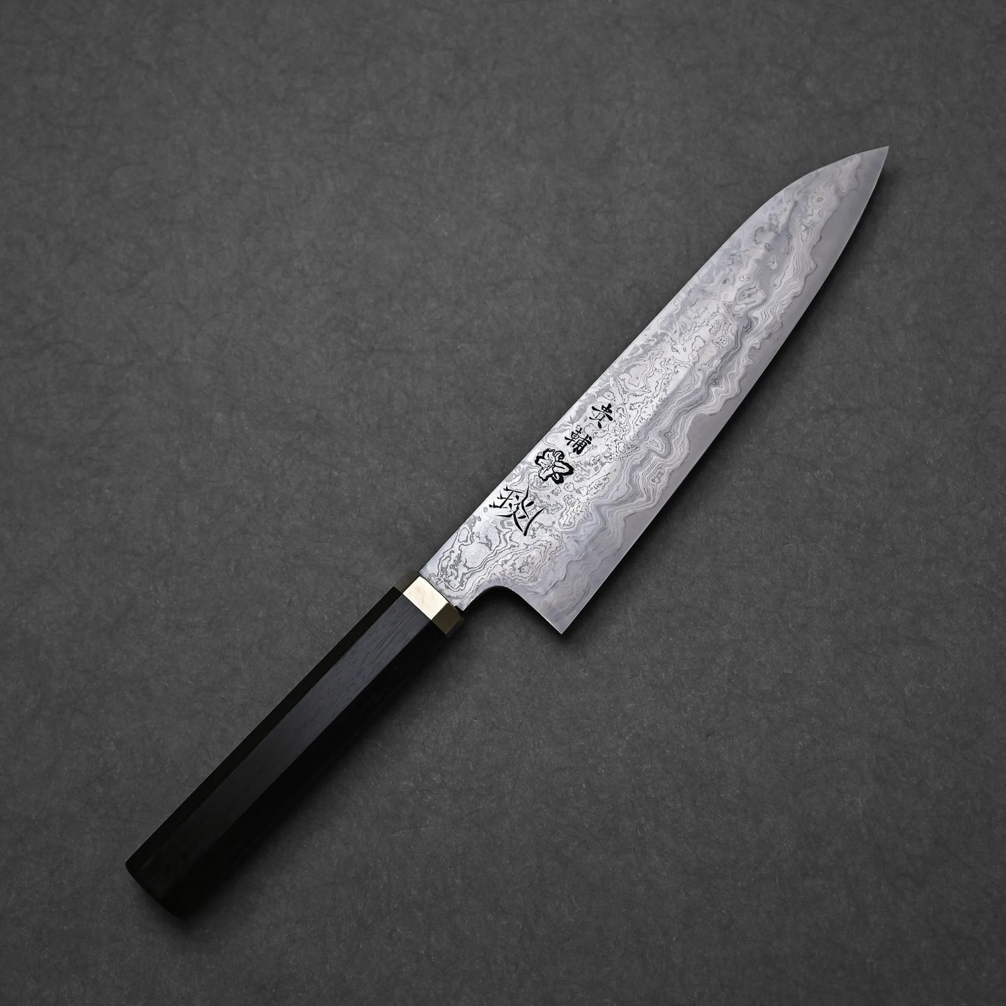 Japanese Damascus steel knife blade with pronounced layered patterns, vertically engraved kanji, and an etched flower motif near the center; dark handle with brass bolster.