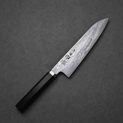 Japanese Damascus steel knife blade with pronounced layered patterns, vertically engraved kanji, and an etched flower motif near the center; dark handle with brass bolster.