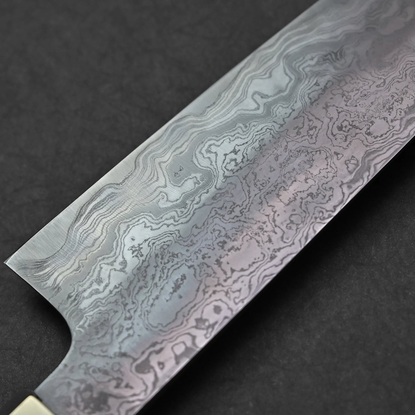 Close-up of a Japanese Damascus steel knife blade featuring layered wavy forge patterns and vertically engraved kanji characters, photographed against a dark textured surface.