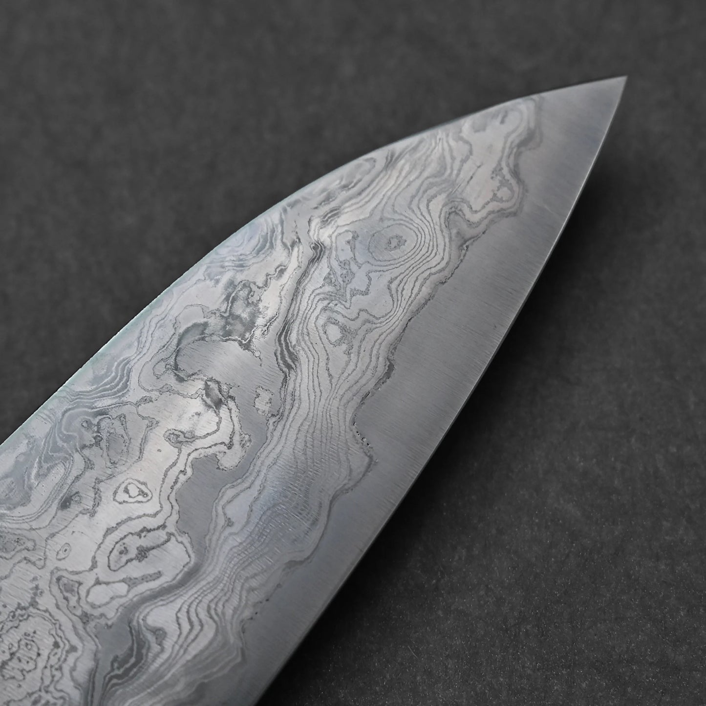 Tip of a Japanese Damascus steel knife blade displaying fine, layered forge patterns and swirling textures characteristic of folded steel craftsmanship.