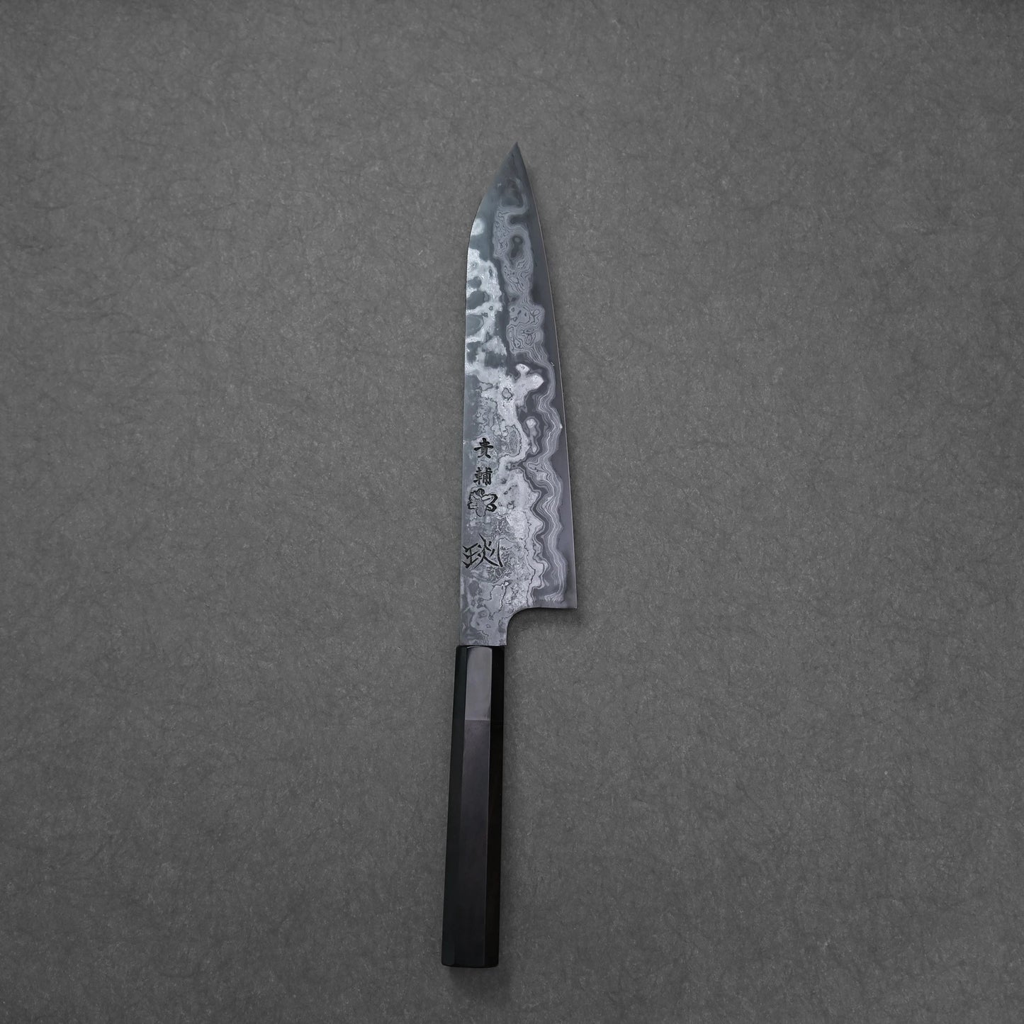 Japanese chef’s knife on a grey textured background, featuring a dark handle and Damascus steel blade with wavy forge patterns and prominent kanji engraving.