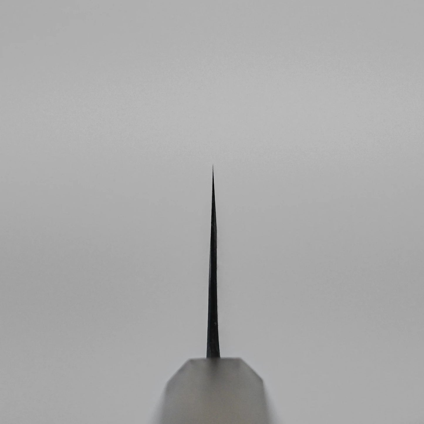 A close-up of the sharp tip of the Kyohei Shindo aogami#2 kurouchi gyuto 240mm is shown front-facing against a plain light grey background. The blade is centered, its edge points up, and it displays an elegant kurouchi finish.