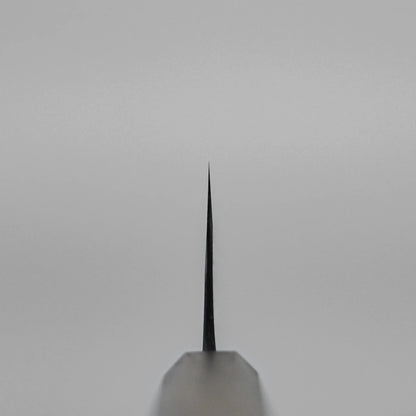 A close-up of the sharp tip of the Kyohei Shindo aogami#2 kurouchi gyuto 240mm is shown front-facing against a plain light grey background. The blade is centered, its edge points up, and it displays an elegant kurouchi finish.